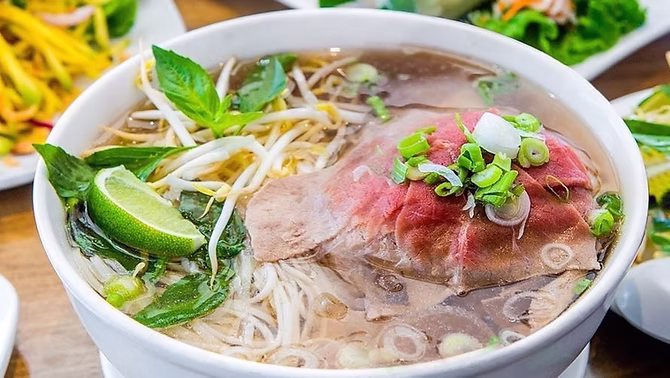 A bowl of traditional Vietnamese Pho with clear simmering broth, rice noodles, fresh herbs, lime, and star anise.